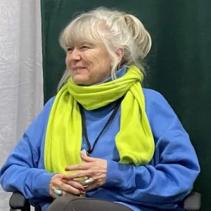 karen is wearing a blue top and bright green scarf and is sitting by a green and white wall